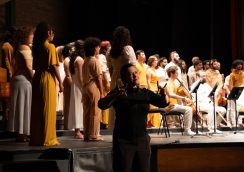 Em um palco, um coral canta ao lado de músicos com instrumentos de corda, enquanto uma intérprete de Libras (Língua Brasileira de Sinais), à frente, traduz a apresentação com gestos expressivos.
