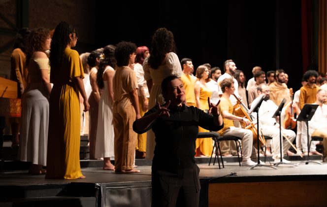 Em um palco, um coral canta ao lado de músicos com instrumentos de corda, enquanto uma intérprete de Libras (Língua Brasileira de Sinais), à frente, traduz a apresentação com gestos expressivos.